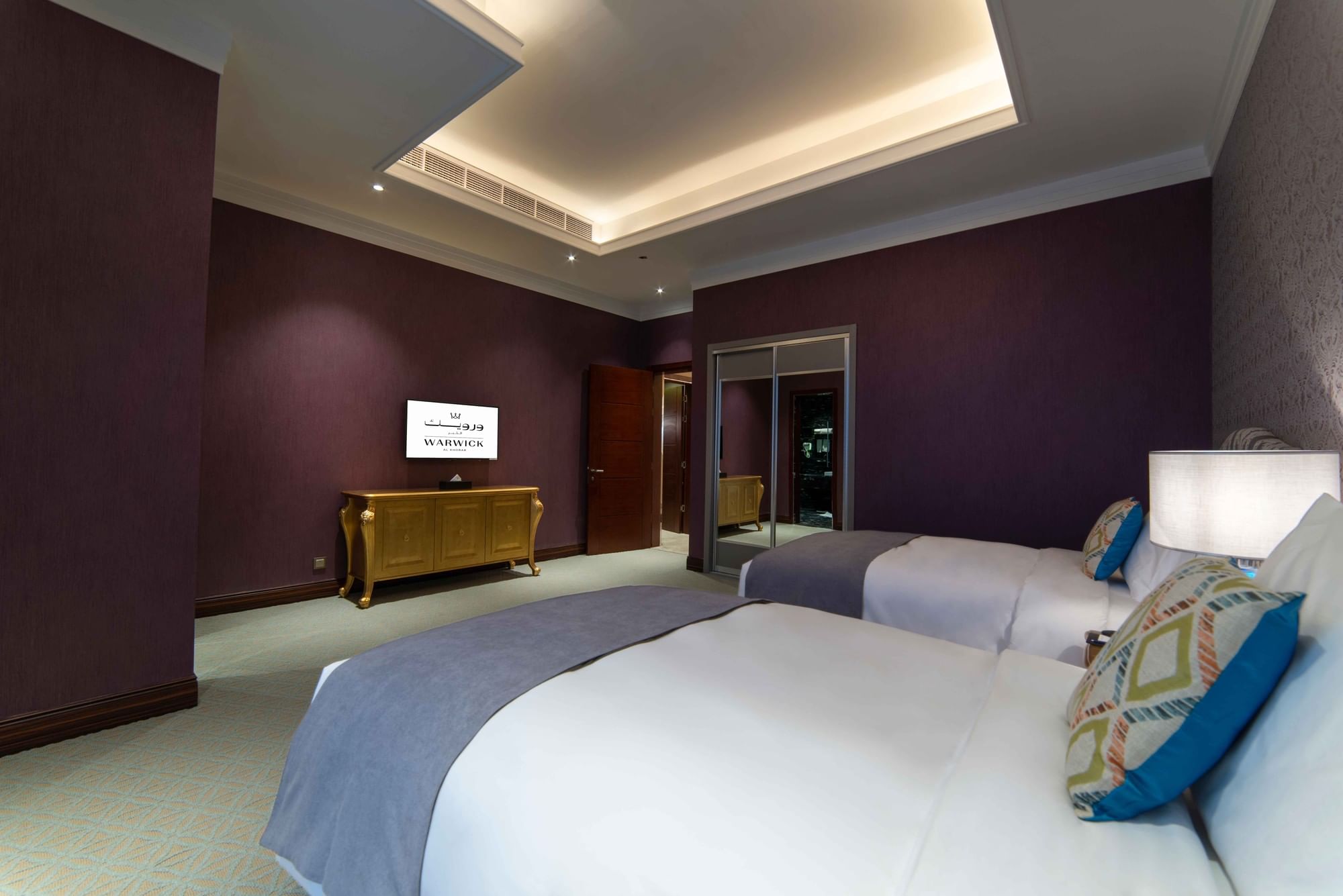 Twin beds with gray throws by a gold-accented dresser and purple textured walls in Penthouse Villa at Warwick Al Khobar