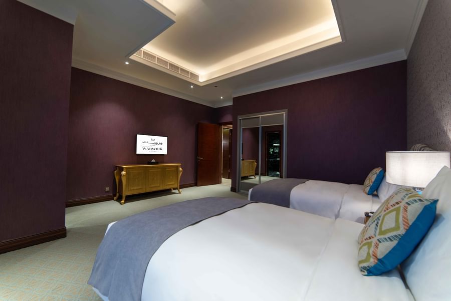 Twin beds with gray throws by a gold-accented dresser and purple textured walls in Penthouse Villa at Warwick Al Khobar