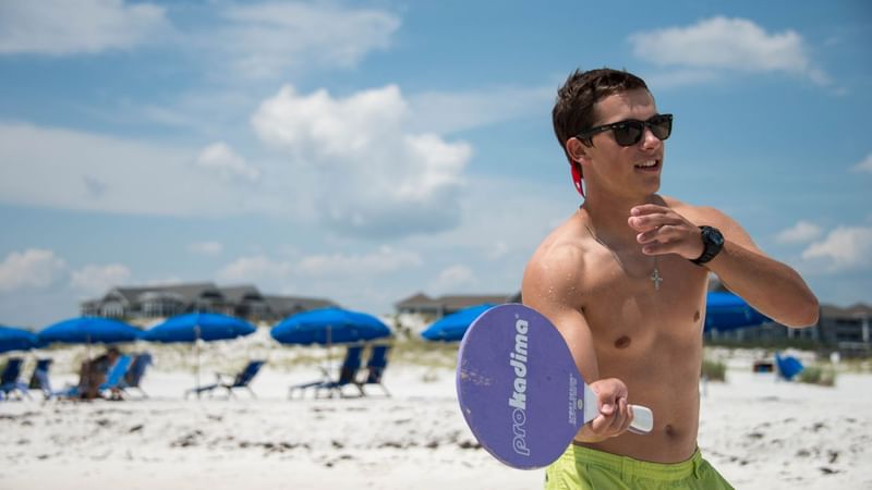 A guy holding paddle rackets on the beach at Watersound Inn
