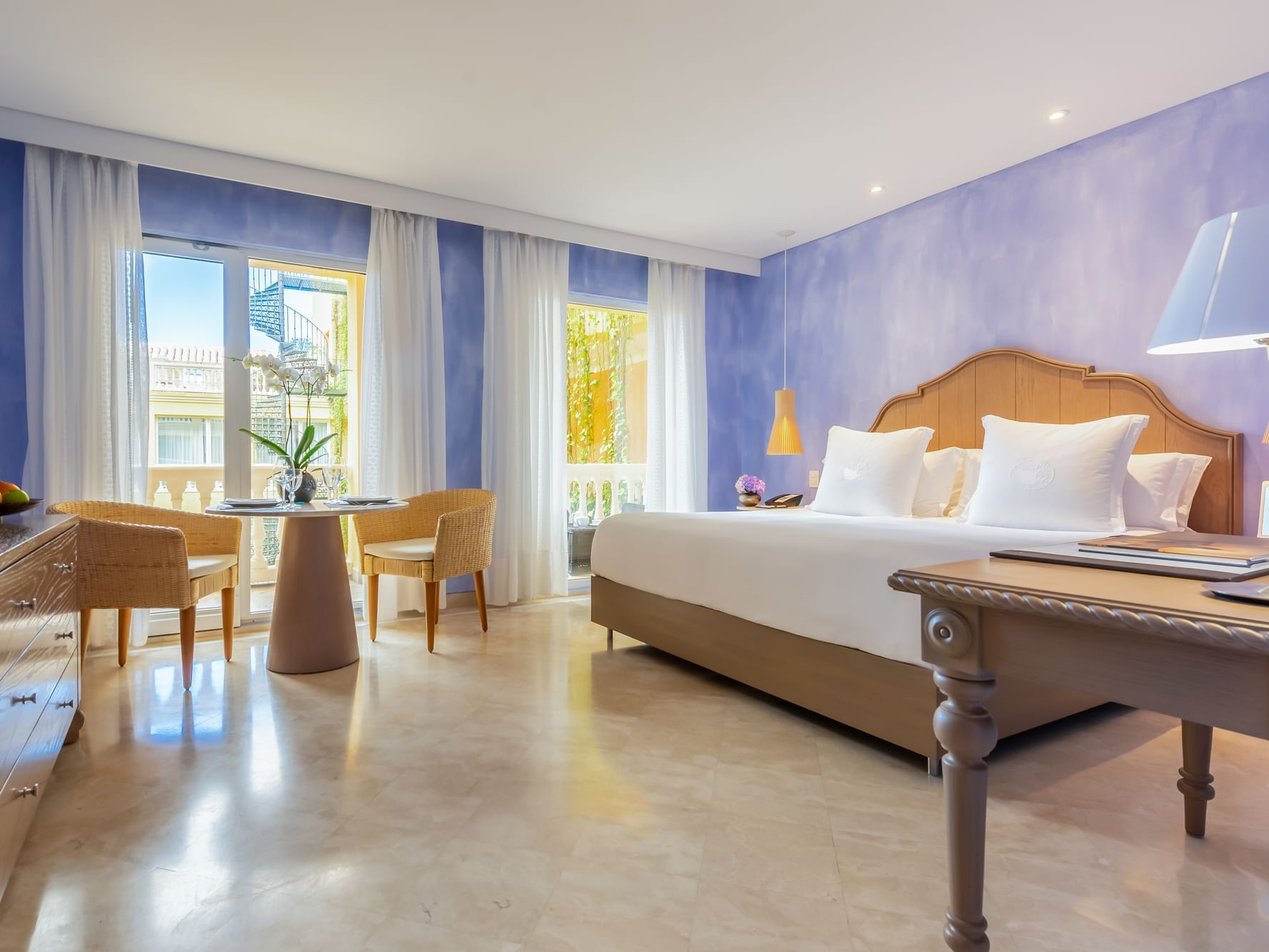 Elegant Premium Balcony room with courtyard view at the Charleston Santa Teresa Hotel in Cartagena. large bed, blue walls, white curtains, round table, and wooden furniture with a lamp.