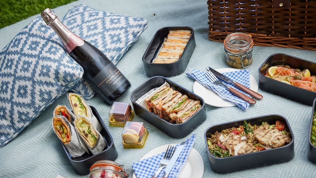 Picnic setup on blanket with sandwiches, salads, olives, cheese, crackers, and a bottle of wine, with a wicker basket nearby.