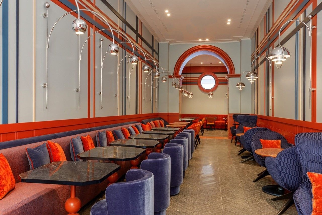 Stylish dining area at the Independent Hotel in Rome with long banquette seating, velvet chairs in deep blue and purple tones, marble tables, red and orange accents, and modern chrome wall lamps under high ceilings.