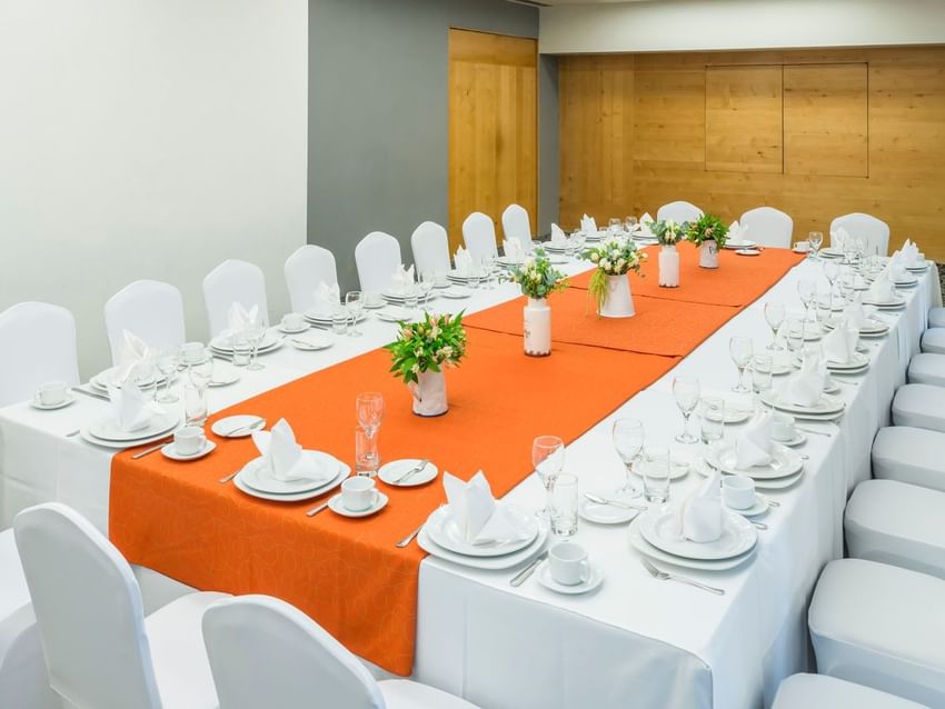 Large conference table in Sidney Venue featuring a bright orange runner and white floral bouquets at Real Inn Perinorte