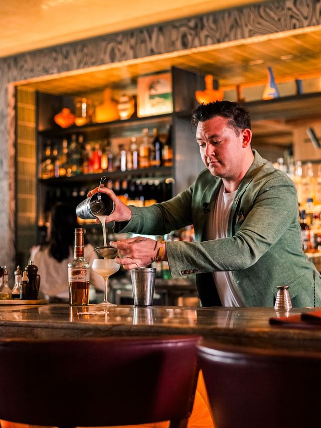 Bartender at Elysium Manly pouring a cocktail from a metal shaker into a glass by a bottle in the Cibaria\'s 55 North Bar