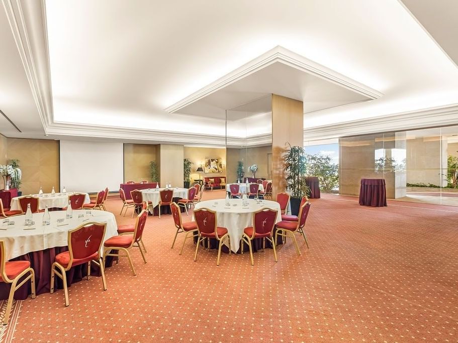 Salón de eventos con mesas redondas y sillas rojas, alfombra roja y plantas en macetas.