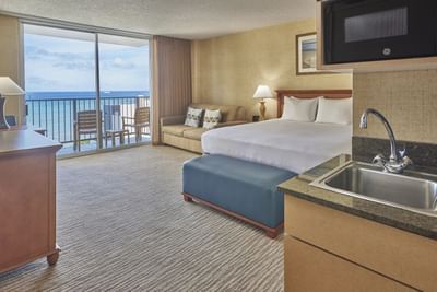 King bed with a footboard bench & cozy loungers by the balcony in Corner Deluxe Ocean View at Waikiki Resort Hotel by Sono