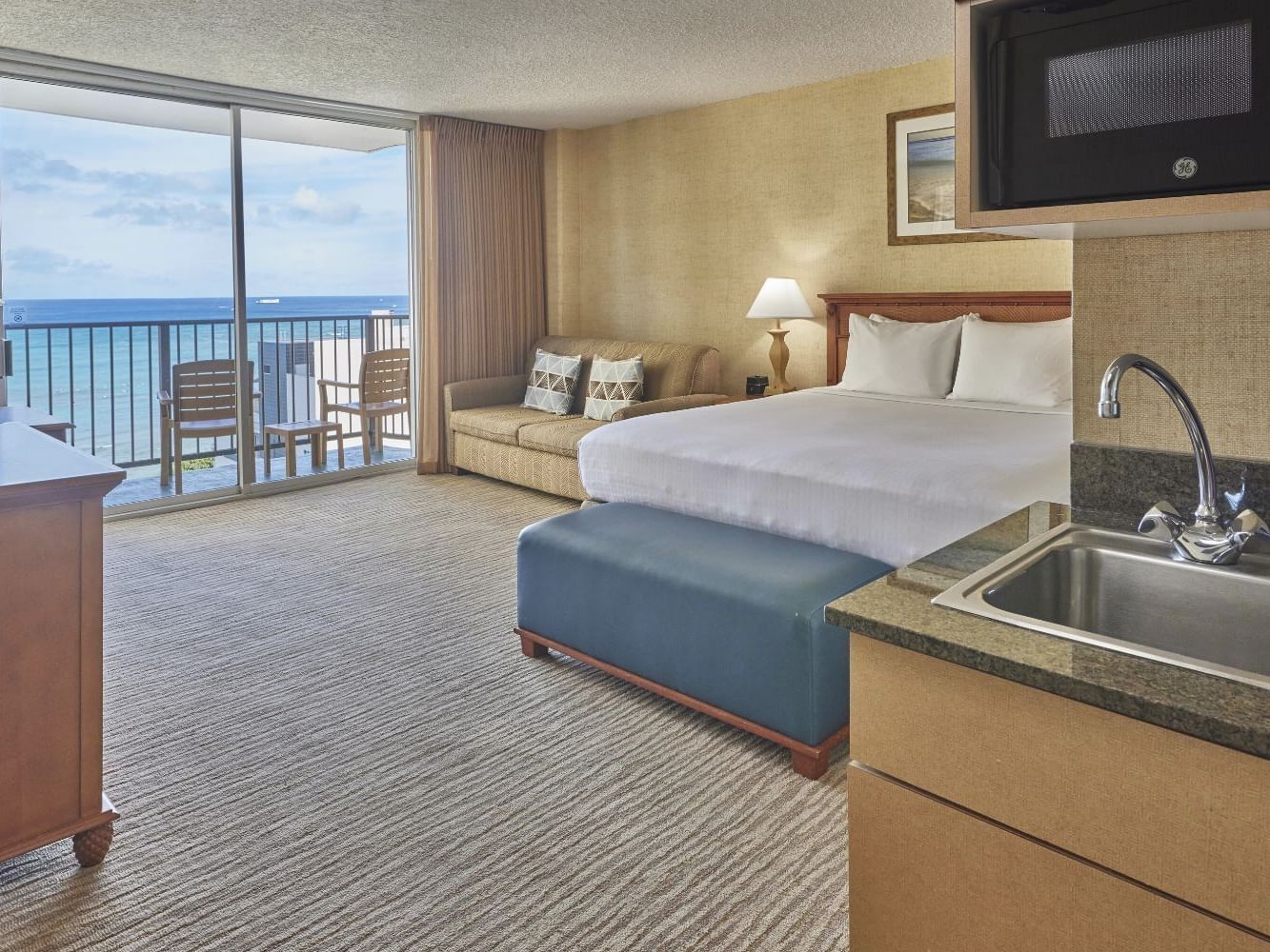 King bed with a footboard bench & cozy loungers by the balcony in Corner Deluxe Ocean View at Waikiki Resort Hotel