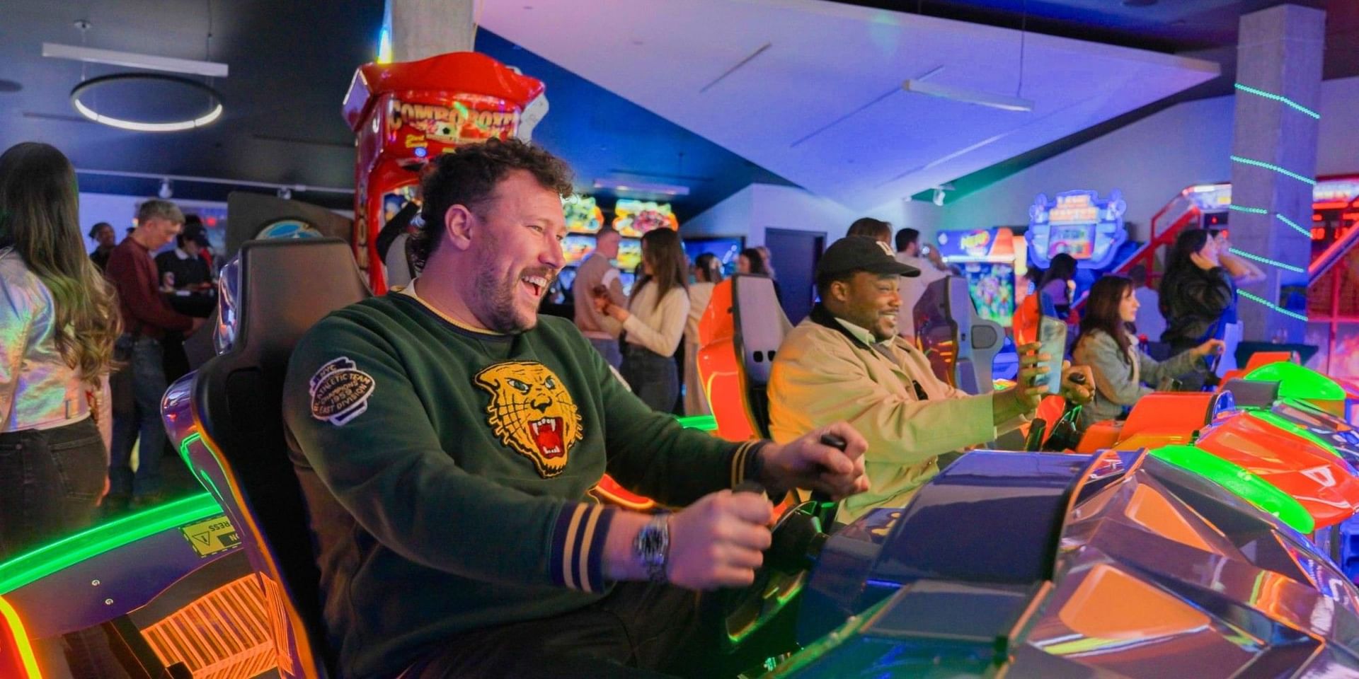 Men laughing while gripping steering wheels in orange racing simulators, under neon lights at Hotel X Toronto