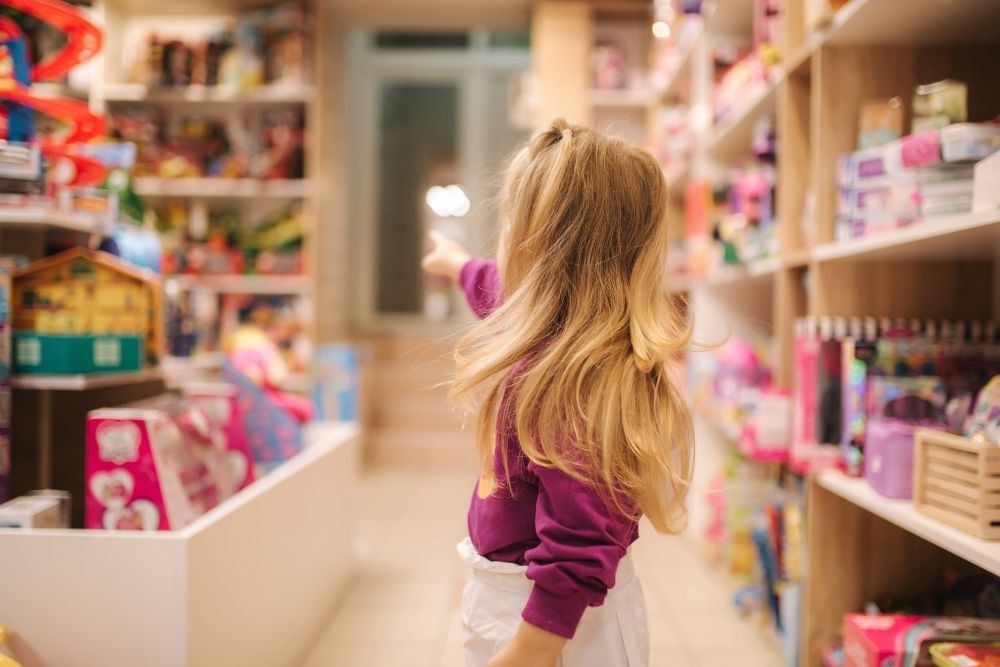 A little girl facing away from the camera points at something on a shelf in an aisle at a toy store. 