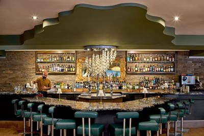 Ampersand 27 bar area with bartender standing behind counter, shelves of liquor bottles, and green stools around the counter.