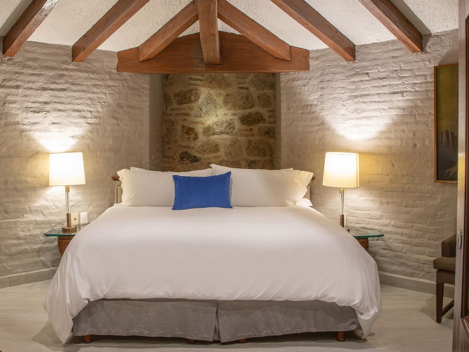 King bed with white linens and blue accent pillow beneath exposed wooden beams and textured stone walls at Quinta Real Guadalajara