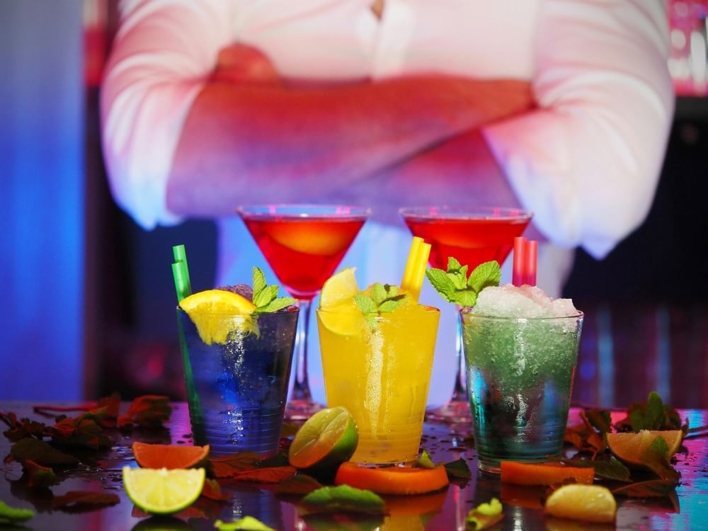 A bartender in a white shirt stands behind an array of colorful cocktails on a bar surrounded by fruit garnishes.