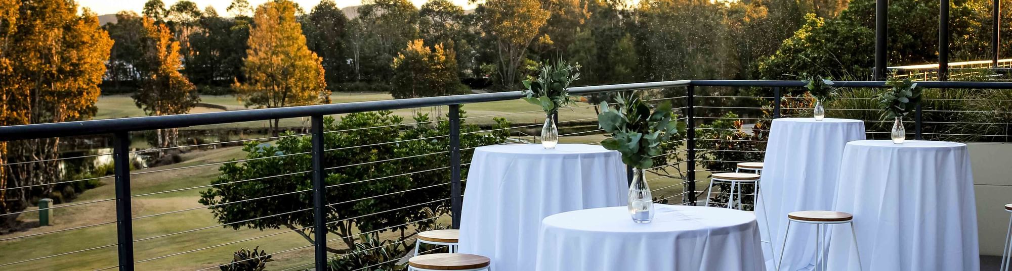 Four round tables with white tablecloths and vases with greenery on a balcony overlooking a garden and trees.