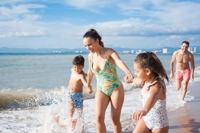 Family splashing and playing together in shallow ocean waves under a partly cloudy sky at Playa Los Arcos Puerto Vallarta