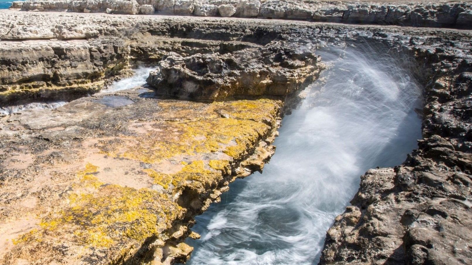 Turquoise water flows through a narrow rocky channel carved into limestone formations at Devil's Bridge under a clear blue sky.