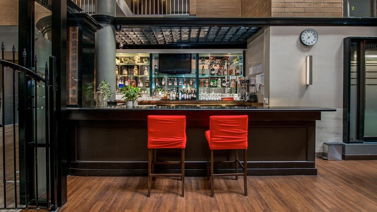 Modern hotel bar with dark wood counter, two red leather stools, and backlit shelving displaying bottles and glassware.