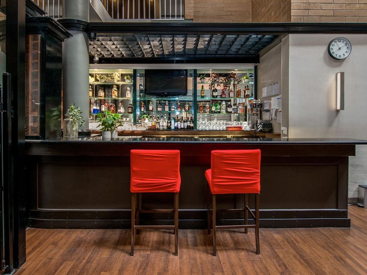 Modern bar with dark wood counter, two red leather stools, and backlit shelving at Coast Calgary University Area Hotel by APA