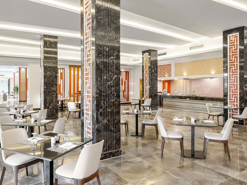 A modern, well-lit restaurant with marble columns, white chairs, and tables set with glassware and napkins.