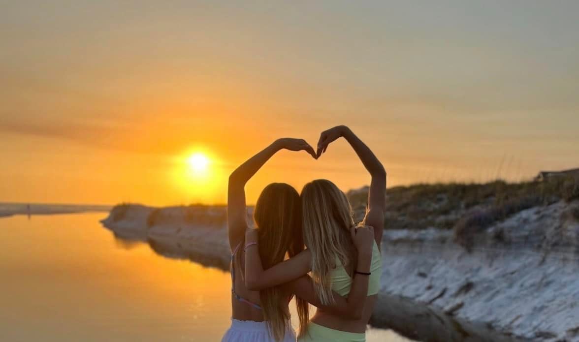 Two women hugging with hands forming a heart shape against a sunset sky and sandy shore.