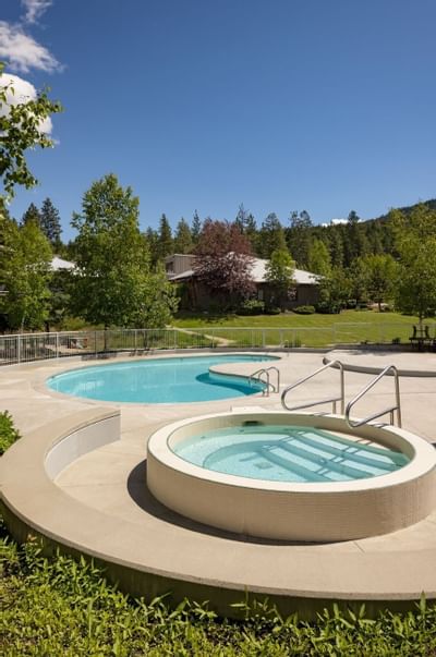 Outdoor pool and hot tub area with surrounding fence, greenery, and a house in the background.