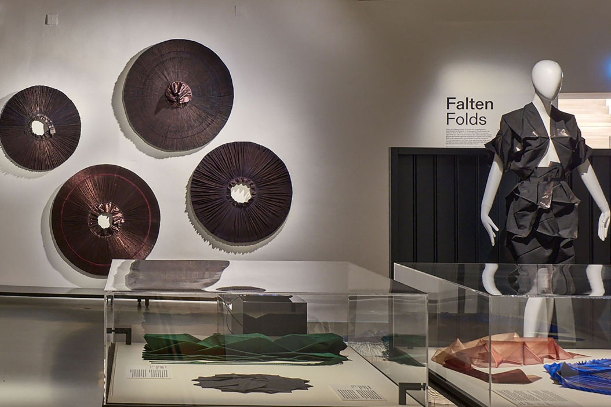 Ausstellungsraum im MAK Wien mit moderner Mode und Designobjekten, darunter ein schwarzes avantgardistisches Kleid und geometrische Installationen zum Thema Falten.