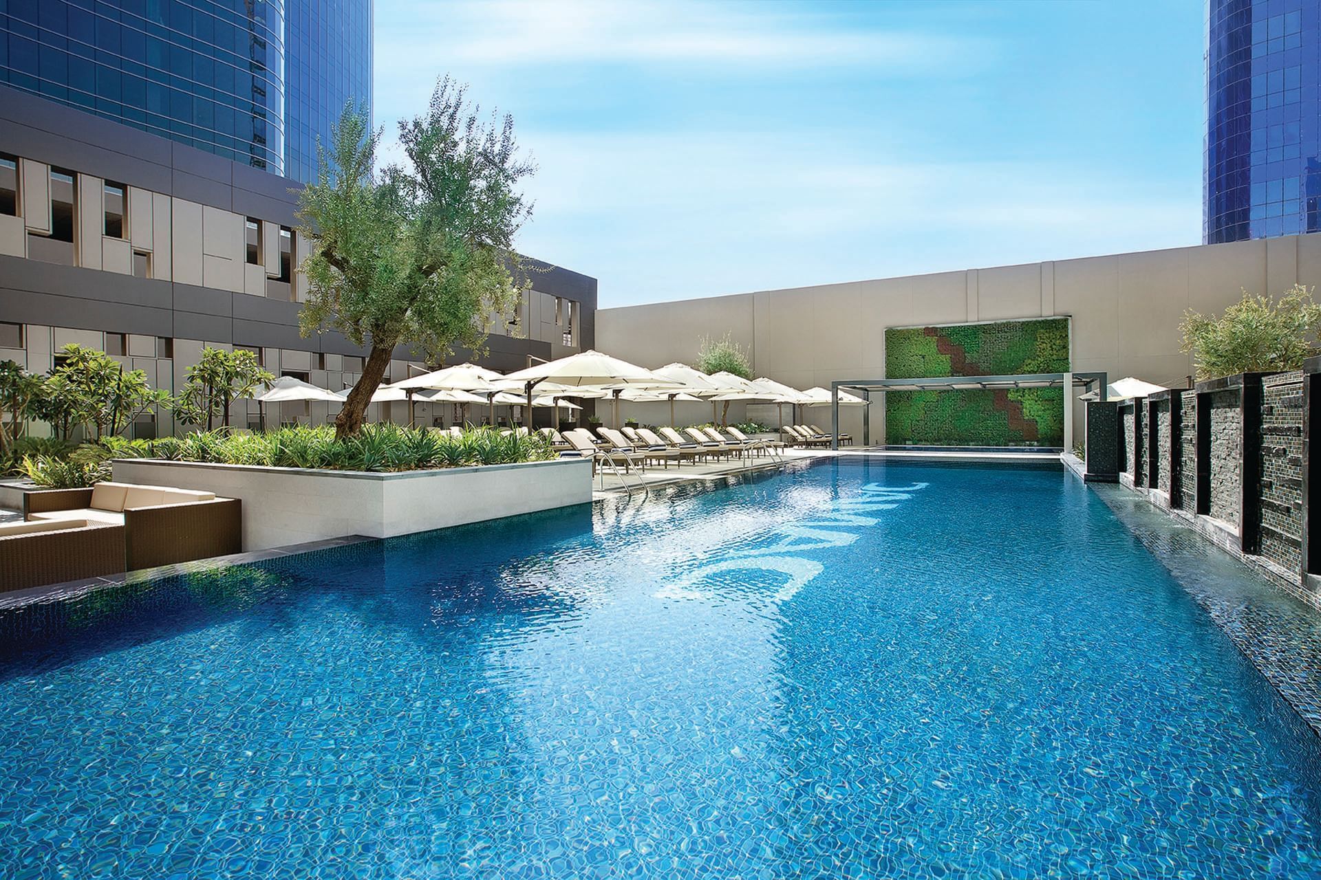 Outdoor pool with lounge chairs under umbrellas, flanked by greenery at DAMAC Maison Cour Jardin Dubai