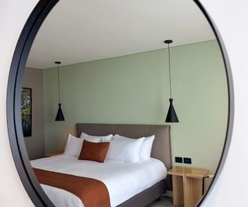 Round mirror reflecting a white bed with rust-colored accents and pendant lights at Unika Hotels, an elite Bogota hotel