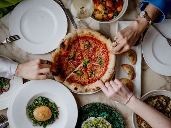 Several hands reaching for a pizza surrounded by various dishes on a marble table at The Kitchens