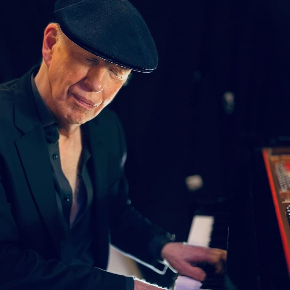 Man in black suit and hat playing the piano with a smile.