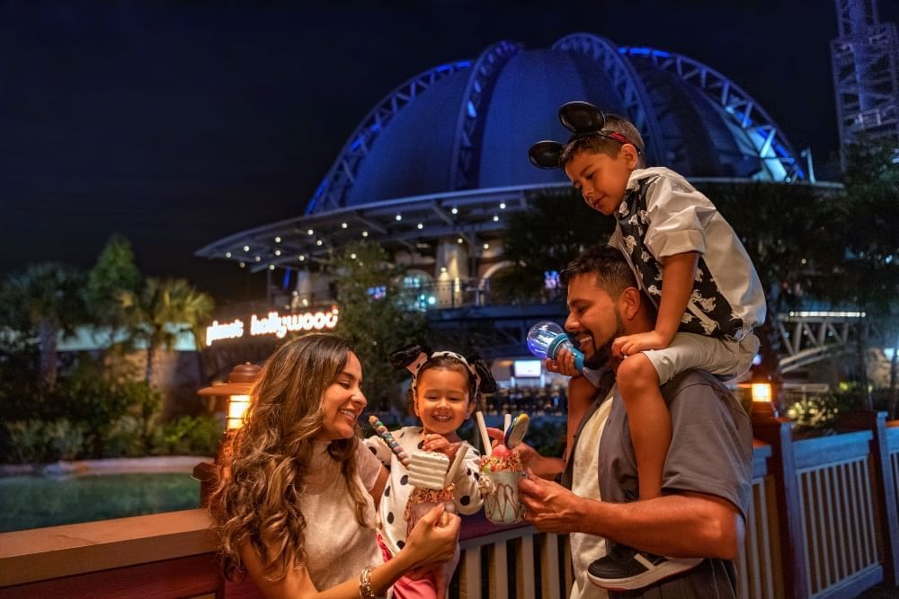 A mom and a dad carrying two children share epic milkshakes standing in front of Planet Hollywood at nighttime.