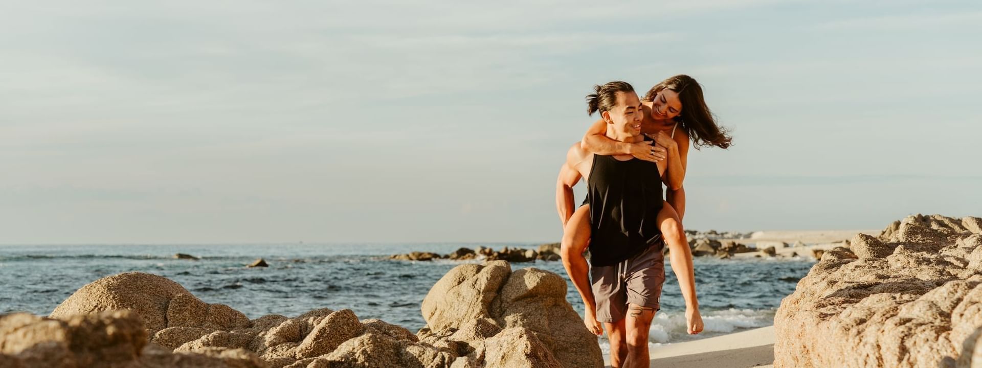 Joyful man giving a piggyback ride to a woman on a rocky beach at sunset, near Marquis Los Cabos Resort