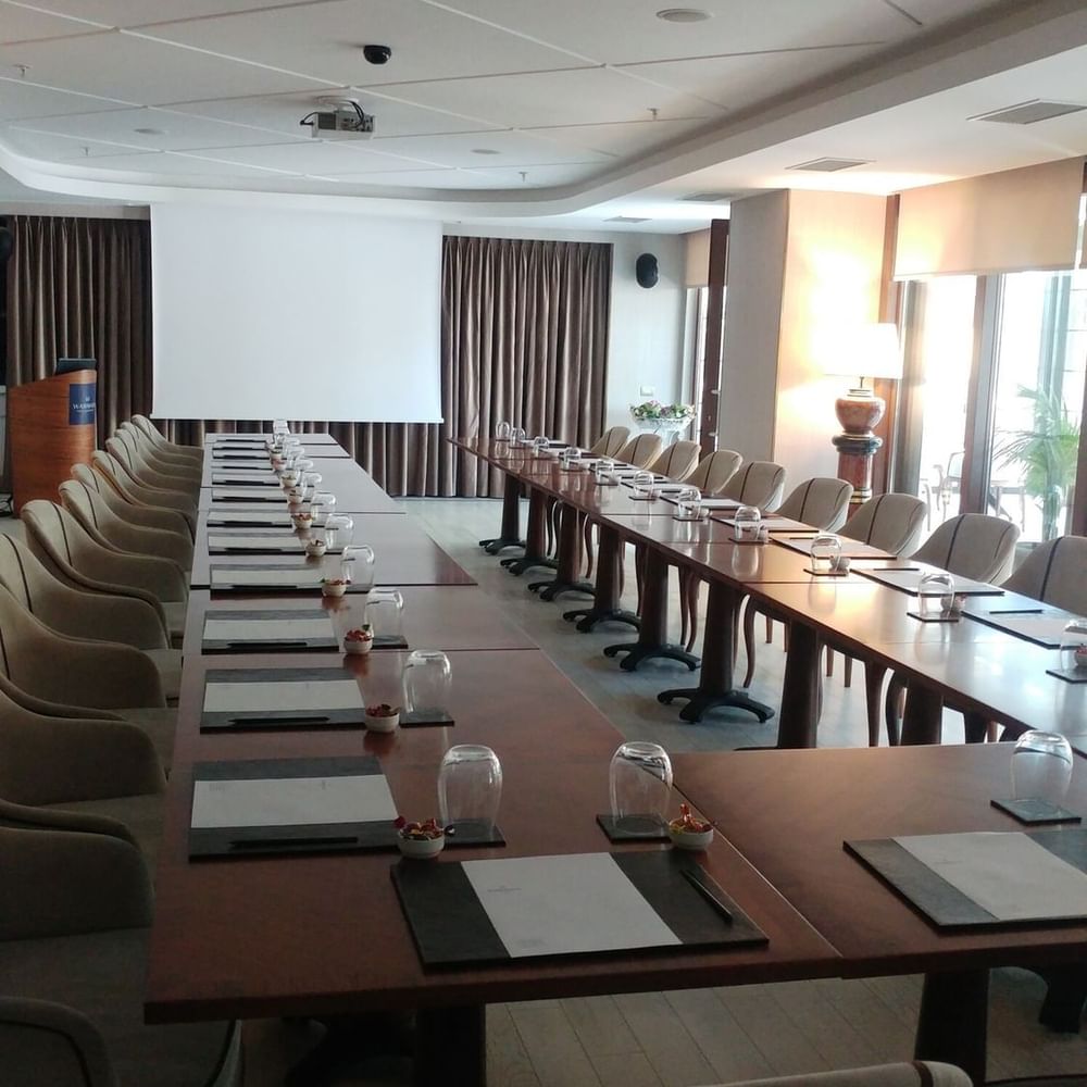 Long table with glasses by plush chairs surrounding a projector screen in Kugu 2 Hall at Warwick Ankara