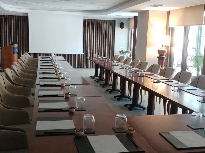 Long table with glasses by plush chairs surrounding a projector screen in Kugu 2 Hall at Warwick Ankara