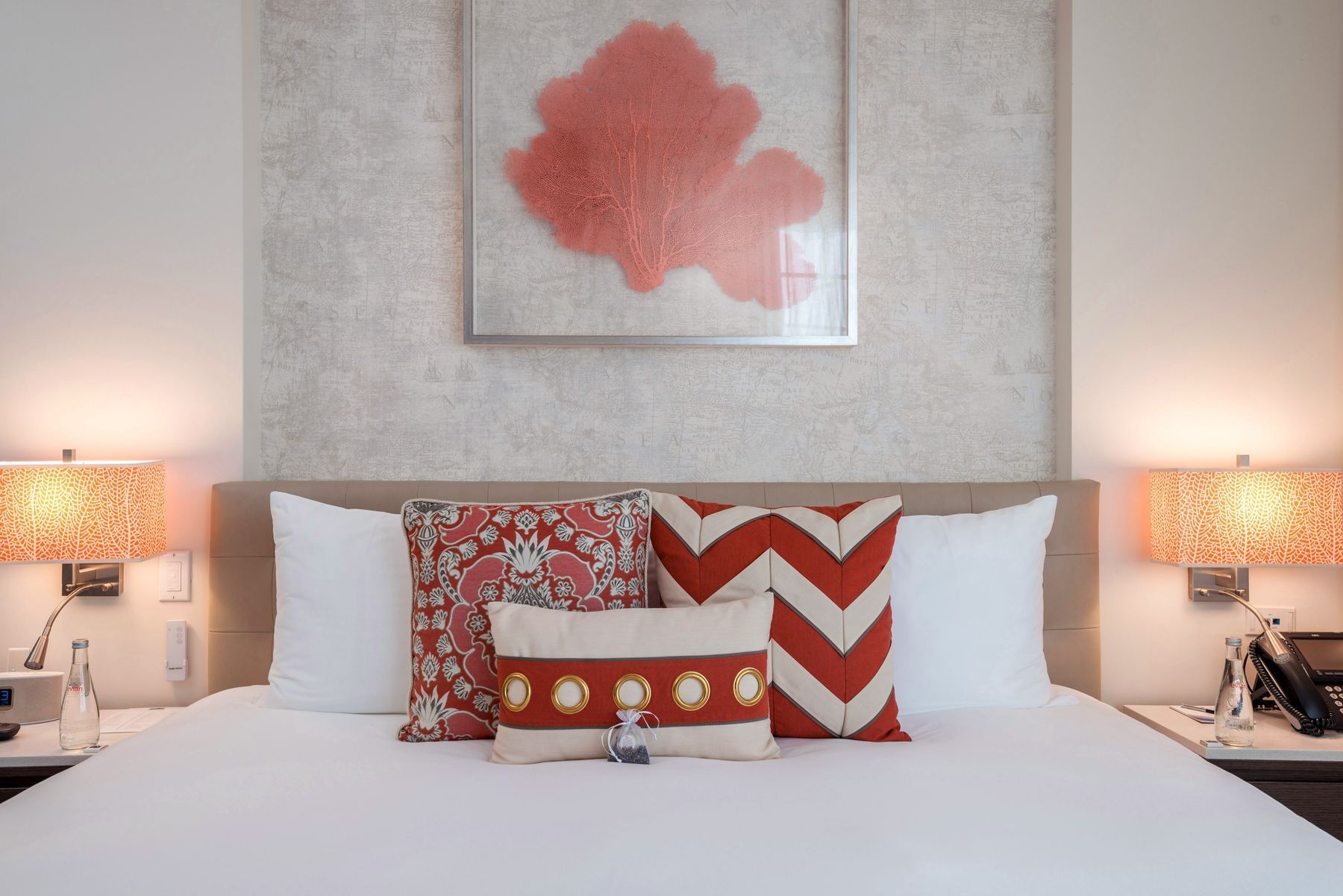 A neatly made bed with white bedding and red and white pillows, with a tan headboard and nightstands with lamps on either side.