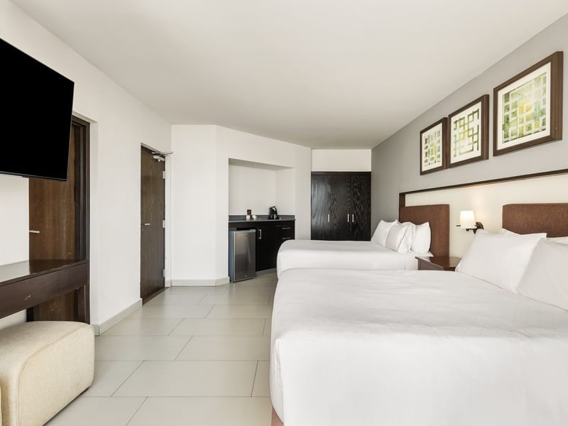 Deluxe room featuring two white plush beds, a wall-mounted TV, and a mini-fridge area at Sunvivia Mazatlán