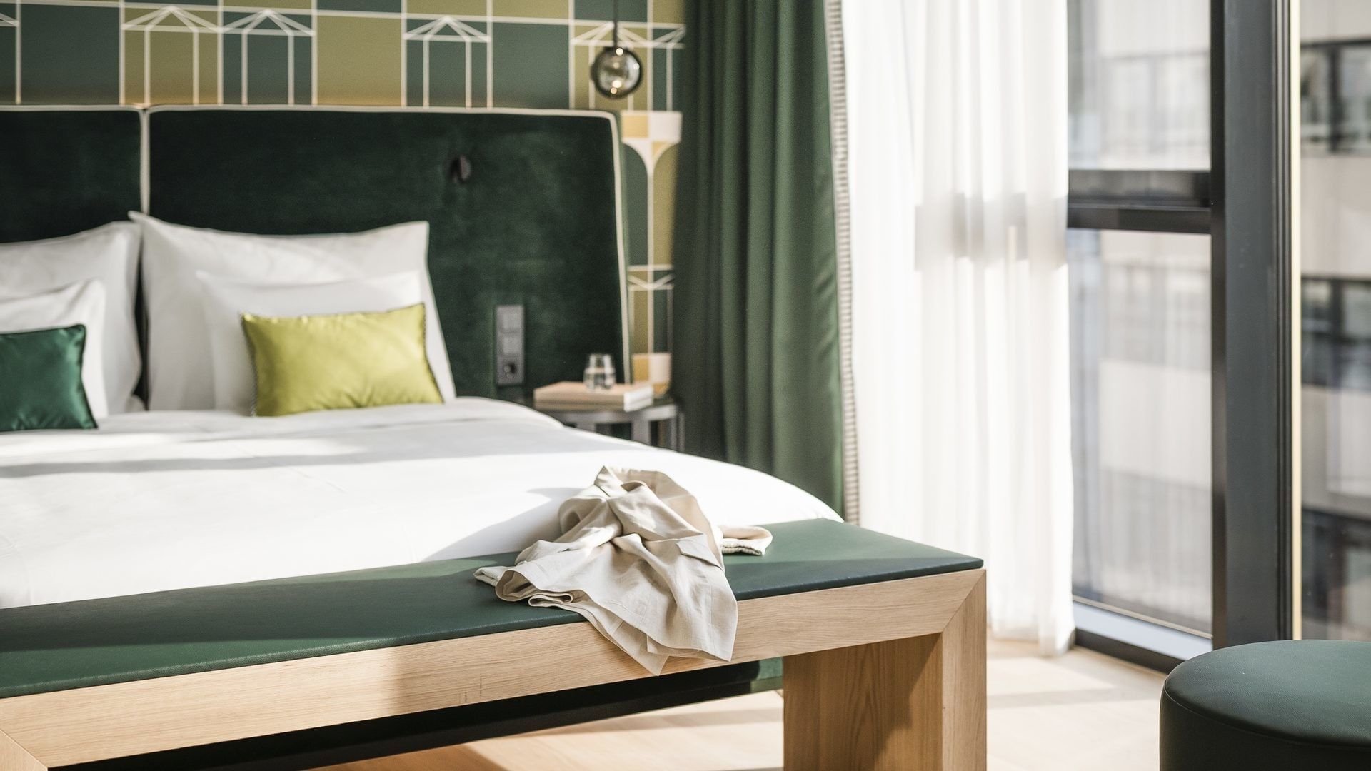 Elegantes Hotelzimmer mit grüner Bettbank, weißen Laken, grünen Kissen und einem Fenster mit Balkon.