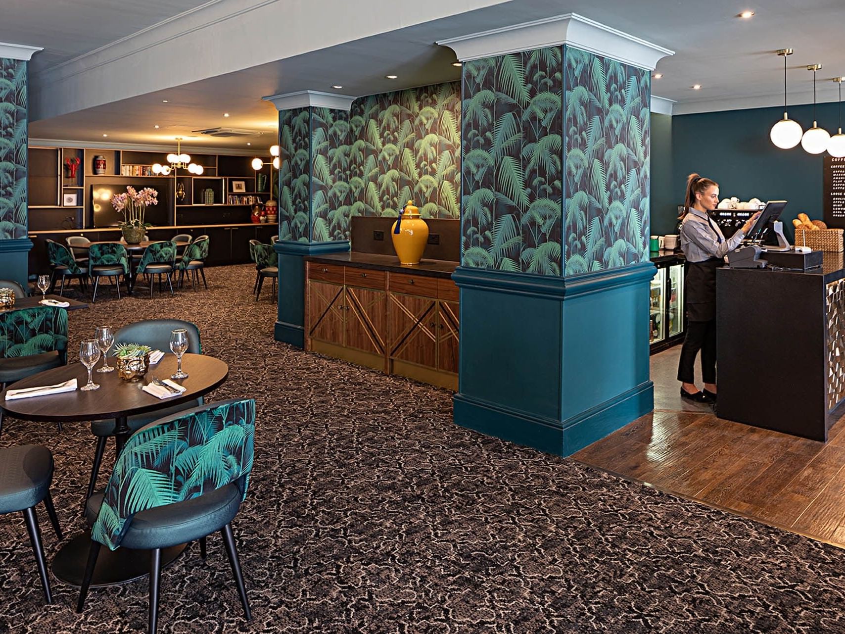Woman standing at a bar counter in a hotel lobby with a tropical pattern on the wall.