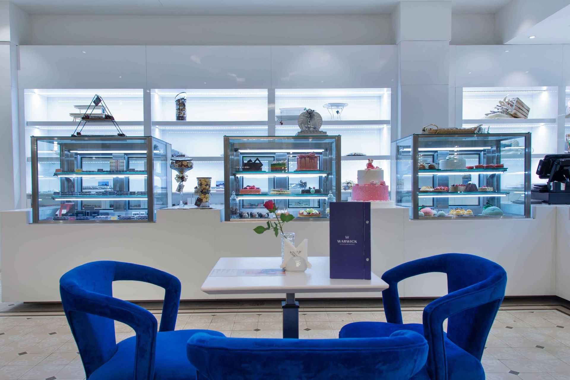 Bright blue armchairs by a small white table in front of glowing glass cake displays in Chocolate Café at Warwick Al Khobar
