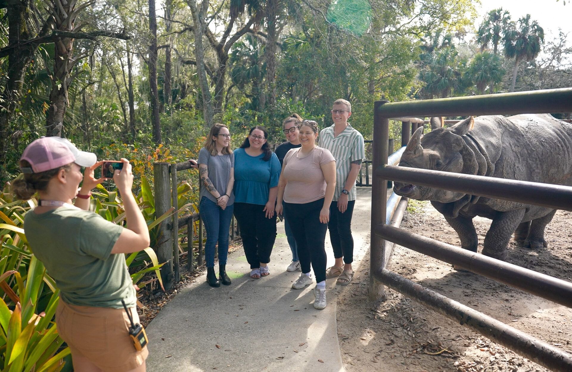 A group of people stand beside a rhino in an enclosure while someone takes their photo.