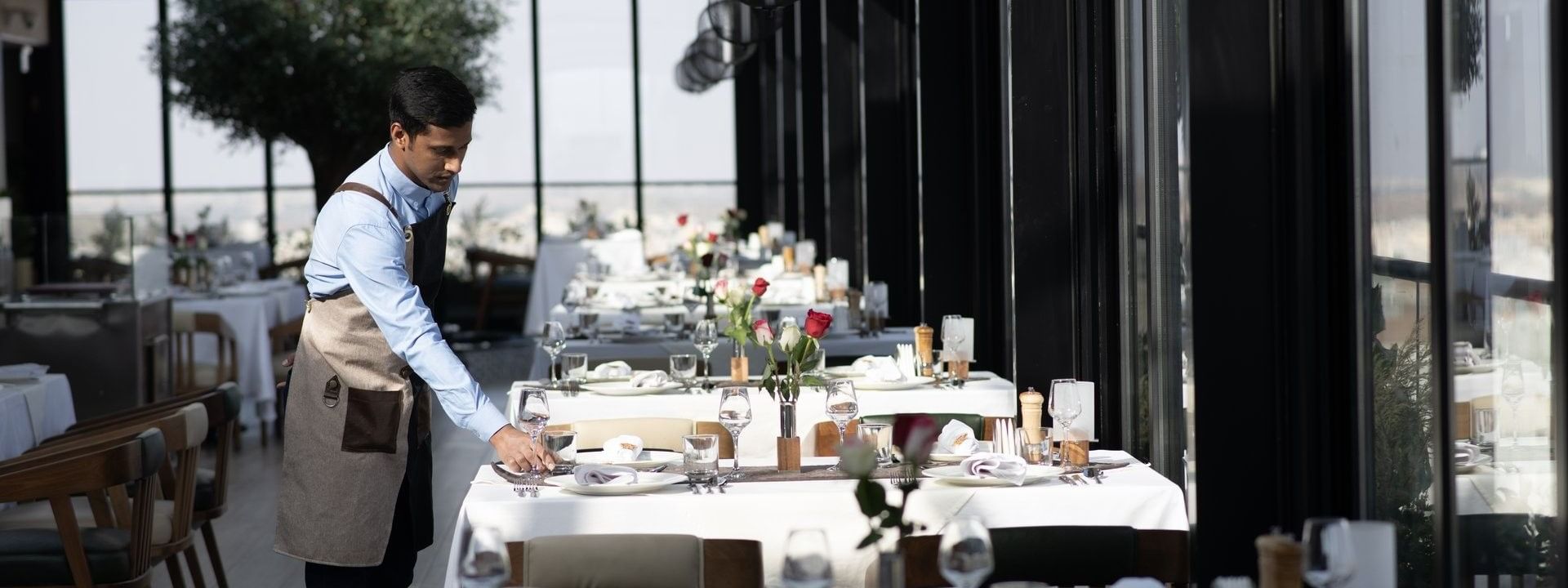 Waiter setting a table by glass walls under black globe pendant lights in Karkan Restaurant at Warwick Al Taif Hotel