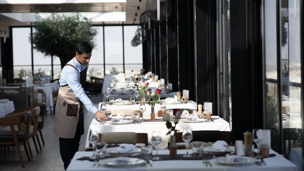 Waiter setting a table by glass walls under black globe pendant lights in Karkan Restaurant at Warwick Al Taif Hotel