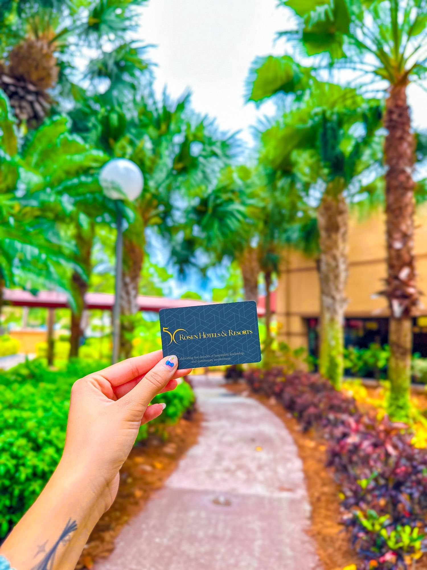 A hand holds a Rosen Hotels and Resorts room key against a backdrop of palm trees and tropical landscaping.  