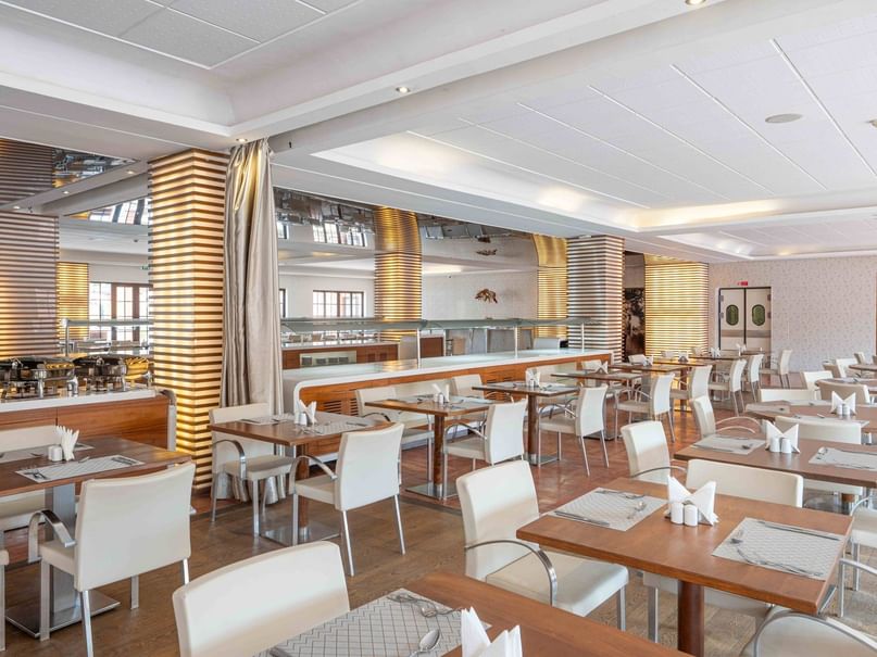 A large restaurant with many wooden tables and white chairs, set in rows, and a buffet counter at the back.