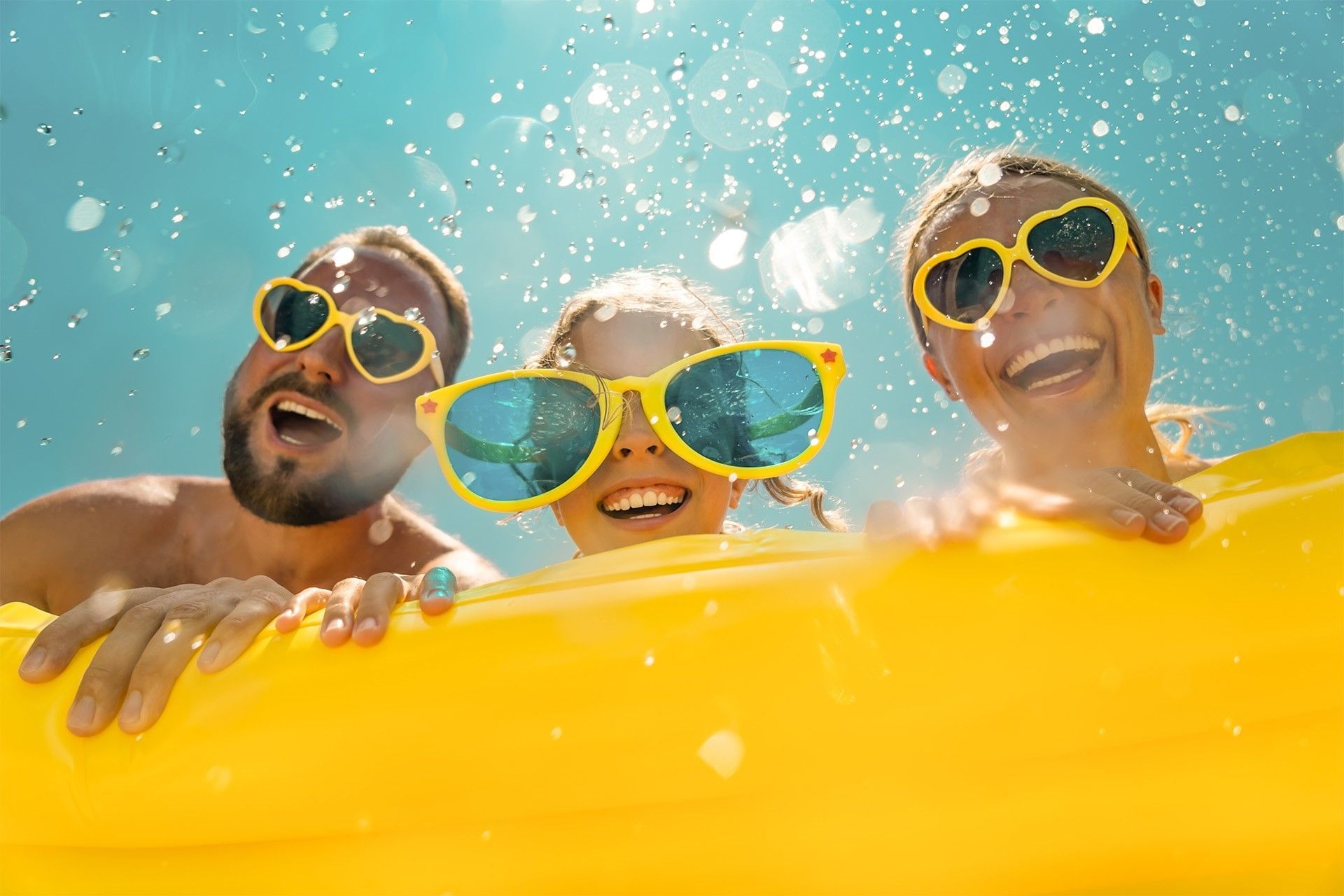 Three people wearing bright yellow sunglasses smile and peak over a large yellow inflatable float in a sunny, blue pool.