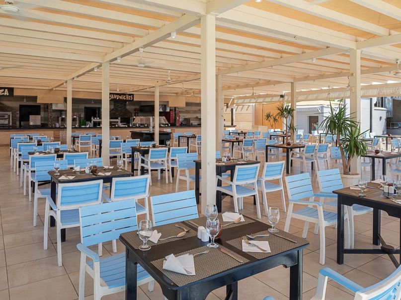 An outdoor restaurant with blue chairs, black tables, and a counter with a pizza sign under a wooden canopy.