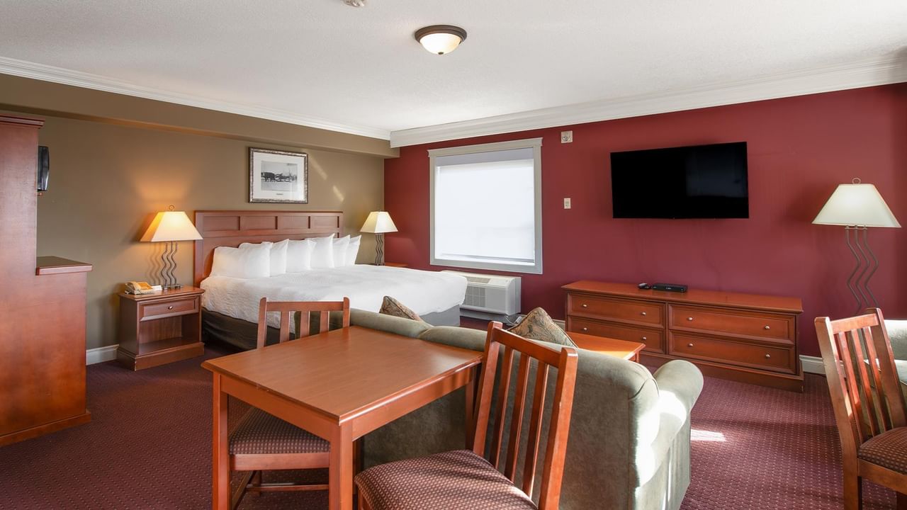 Spacious hotel suite with king bed, dining table, sofa, wall-mounted TV, and burgundy accent wall at Coast Edson Hotel.