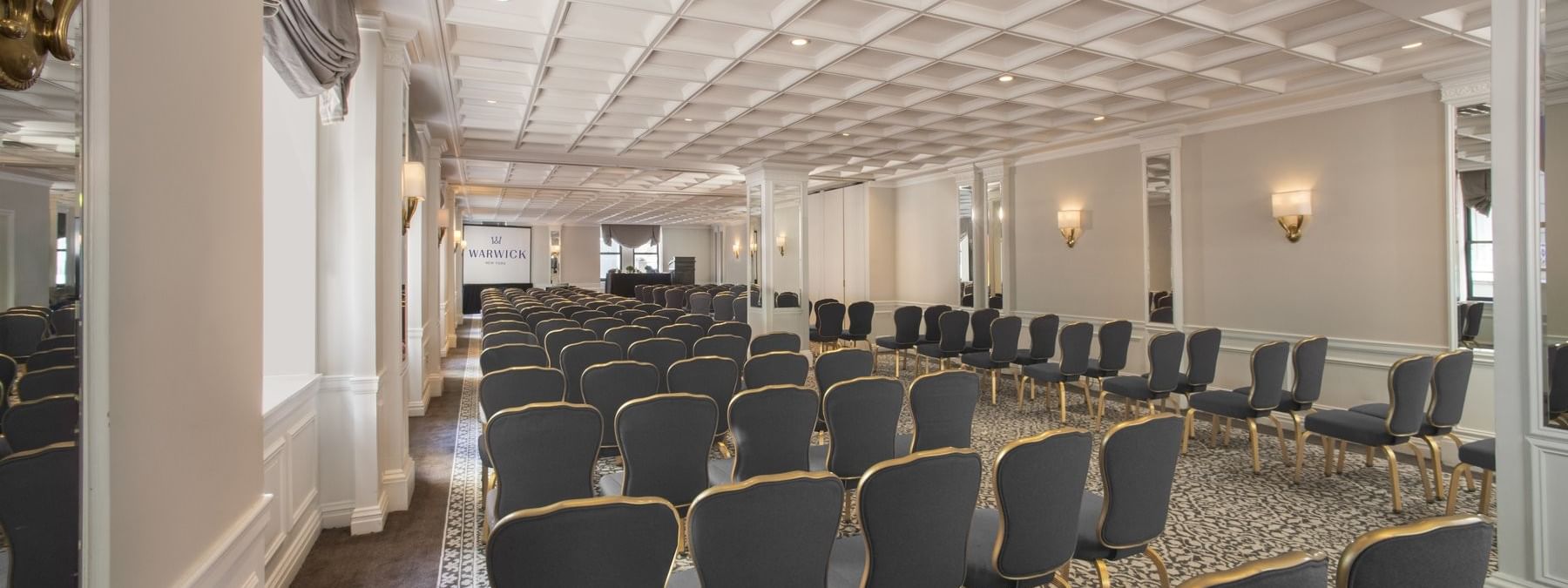 Rows of gray chairs facing a projection screen under a coffered ceiling in Kent/ Surrey/ Oxford at Warwick New York