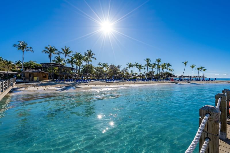 Sunlit tropical beach with calm turquoise water, sandy shores, palm trees, and lounge chairs near El Conquistador Resort