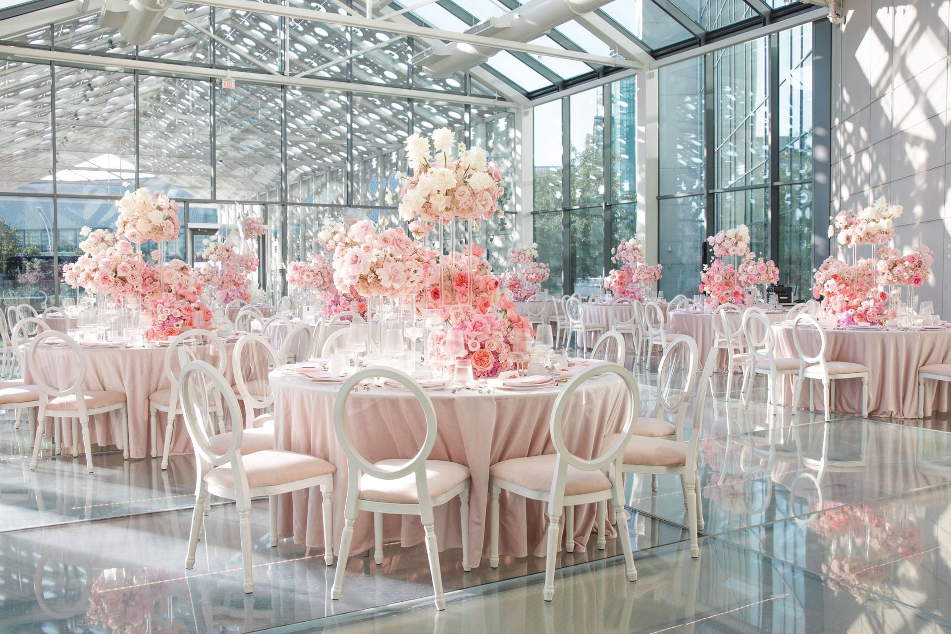 Sunlit glass pavilion filled with round tables and white chairs beside pink floral centerpieces inside Hotel X Toronto