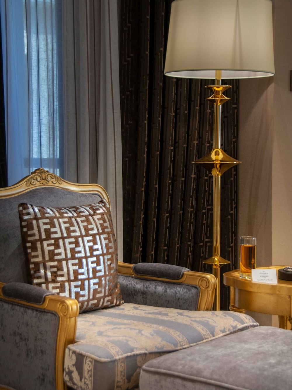 Gold-trimmed chair with a patterned pillow by a gold lamp under dark drapes in Penthouse Villa at Warwick Al Khobar
